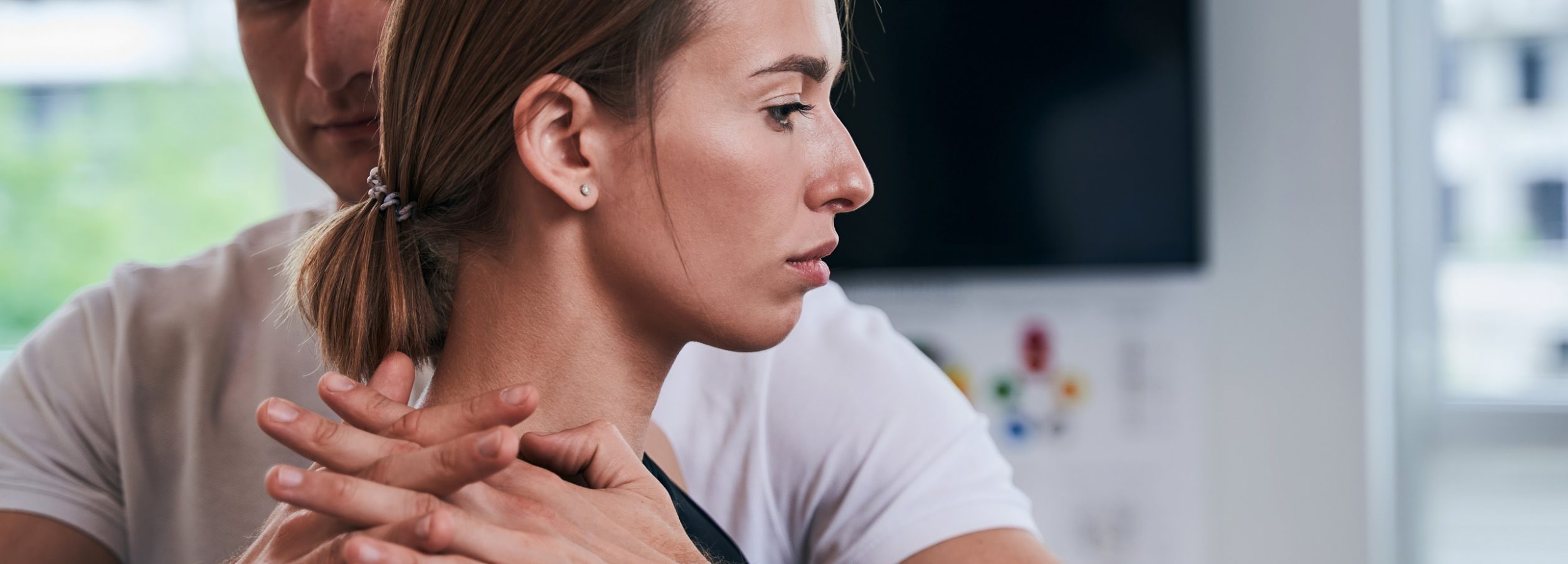 Close up side view portrait of beautiful young Caucasian female standing in room indoors while man physiotherapist doing massage on her shoulder in wellness center