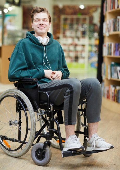 Smiling satisfied handsome young student in casual clothing wearing headphones on neck sitting in wheelchair and looking at camera in modern library